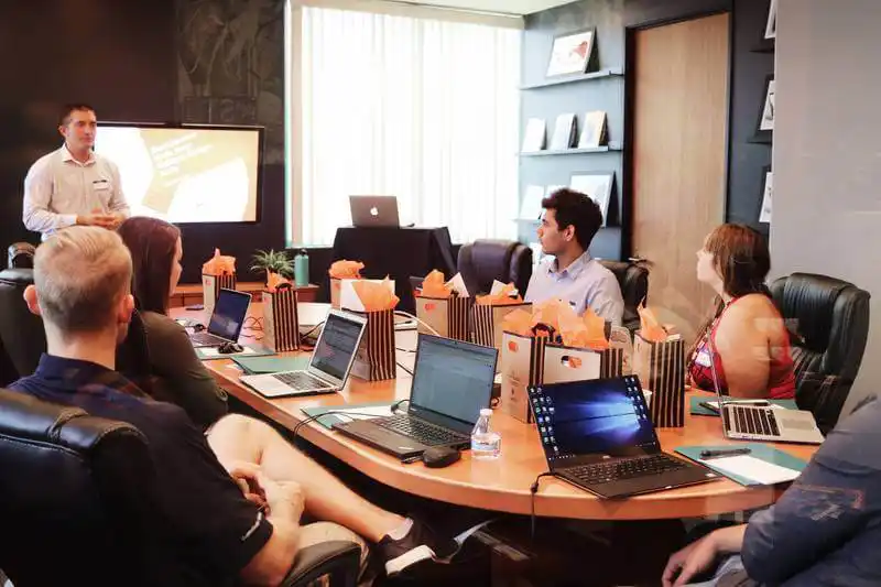 The image shows a group of people in a work meeting. A man is presenting while the otheres are sat around a table looking at him.