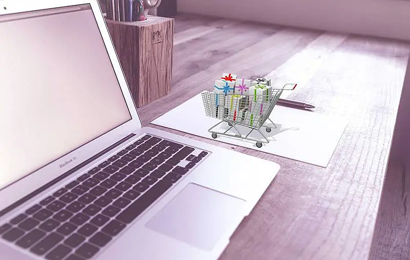 The image shows a laptop on a table, and on its right side there is a mini shopping cart full of wrapped gifts