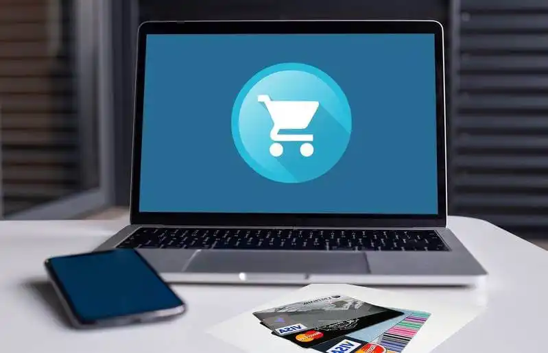 The image shows a laptop on a table, in the screen there is a cart icon. On the table, near the laptop, there is a smartphone ans several credit cards.