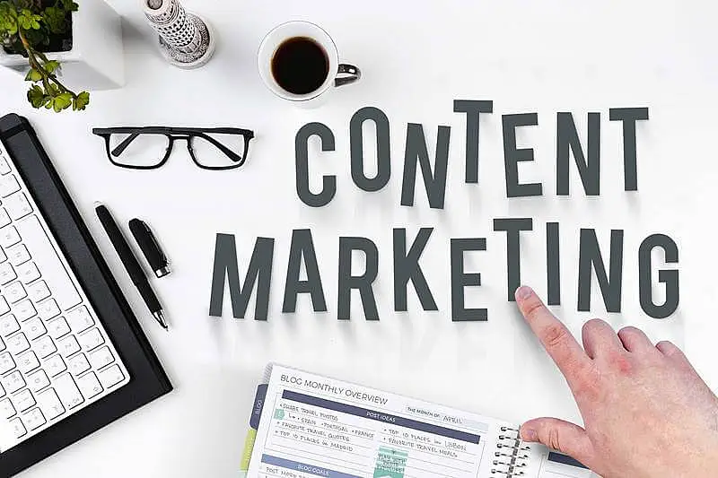 In the image it's writen "Content Marketing" on a table, and around it there us a glass, a mug, a pen, a keyboard, and a notebook.