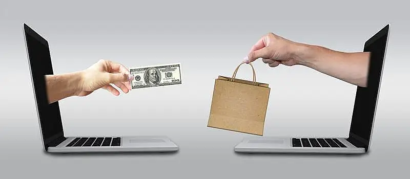 The image shows two hands going out from two laptop screens, one hand is holding a dollar bill and the other one a shopping bag.