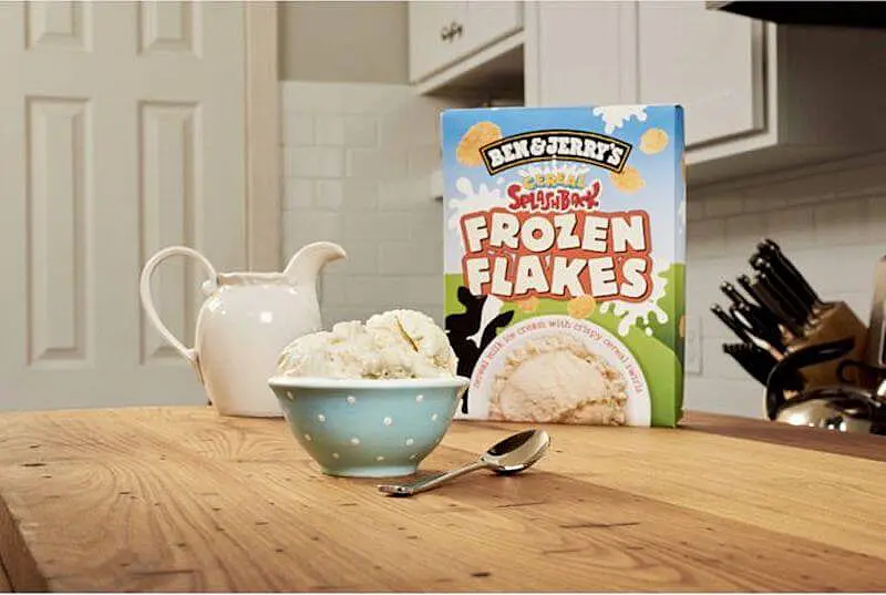 The image shows Ben&Jerry icecream package on a table with a bowl of ice cream in front of it and a sponn, on the side there is a jar of milk. 
