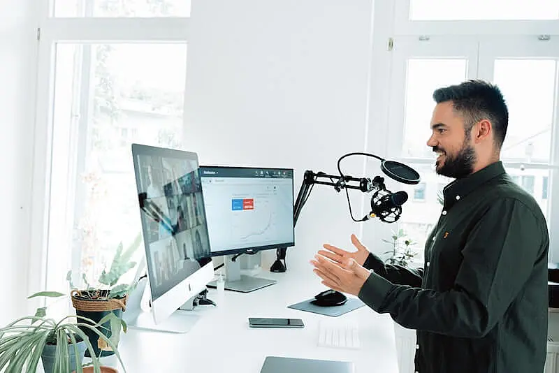 The image shows a man sat in front of 2 screens, he has a microphone installed on the right side of the table.