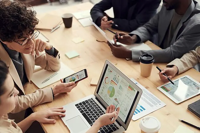 The image shows 5 people in a meeting sitting on a table. Two of them are looking at charts in a computers, one is looking at a tablet, and the other two are looking at a paper.