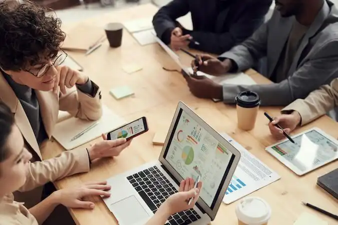The image shows 5 people in a meeting sitting on a table. Two of them are looking at charts in a computers, one is looking at a tablet, and the other two are looking at a paper.