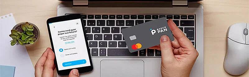 Foto de um notebook na mesa e uma mão segurando um celular e a outra mão segurando um cartão do banco Pan