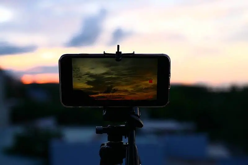 The image shows a smartphone attached to a phone tripod, recording the sunset.