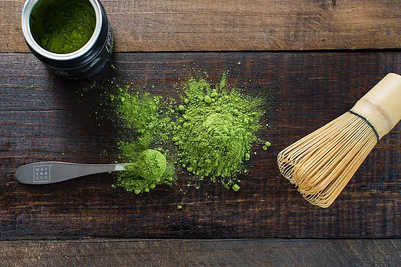 The image shows a can of matcha, a spoon with matcha, and there is also the matcha powder on the table.