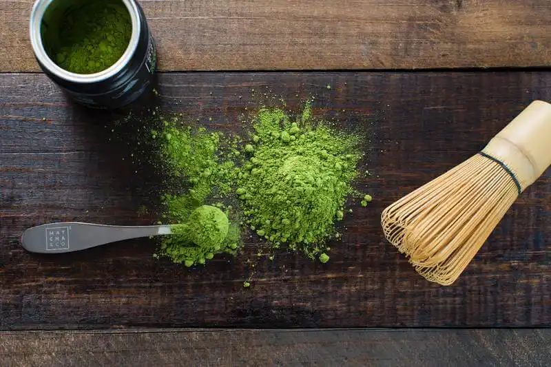 The image shows a can of matcha, a spoon with matcha, and there is also the matcha powder on the table.
