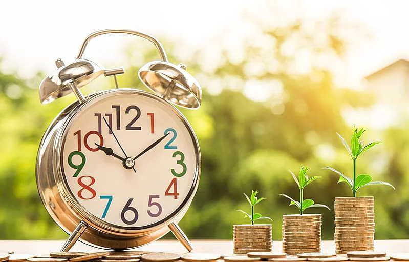 The image shows a clock and 3 towers of coins with a plant on the top of each tower.