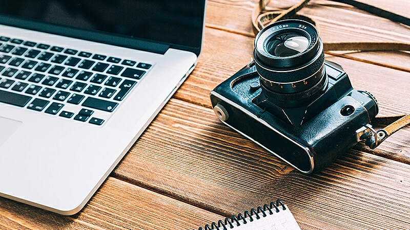 Foto de um notebook, caderno e câmera em cima da mesa