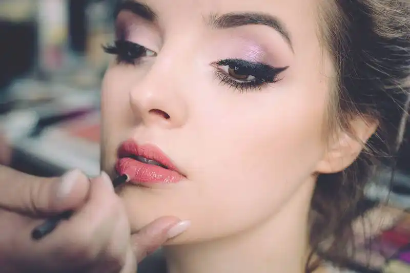 The image shows a woman's face with makeup on, someone is passing lipstick on her.