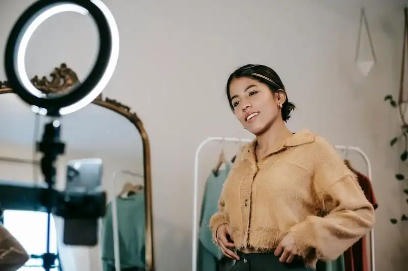 The image shows a girl recording a video in front of a ring light and a smartphone.