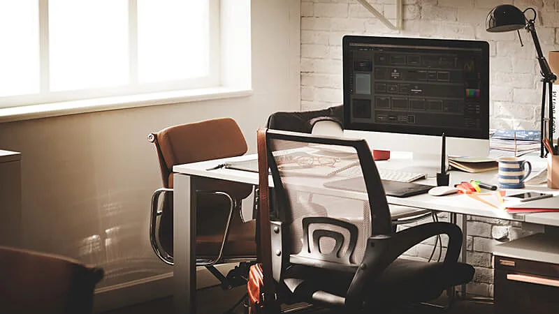 imagem que mostra um escritório em casa com uma mesa cheia de dispositivos e uma cadeira próximo a duas janelas