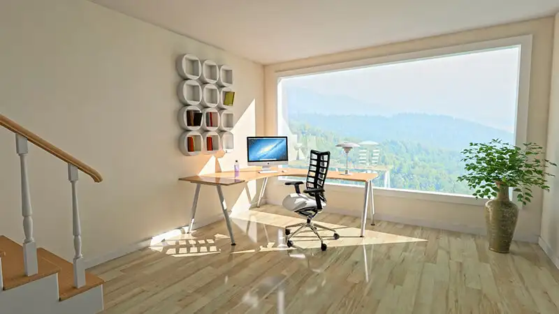 imagem que mostra uma mesa e uma cadeira de escritório em uma sala extensa com uma janela grande, chão de madeira e um vaso de plantas na direita