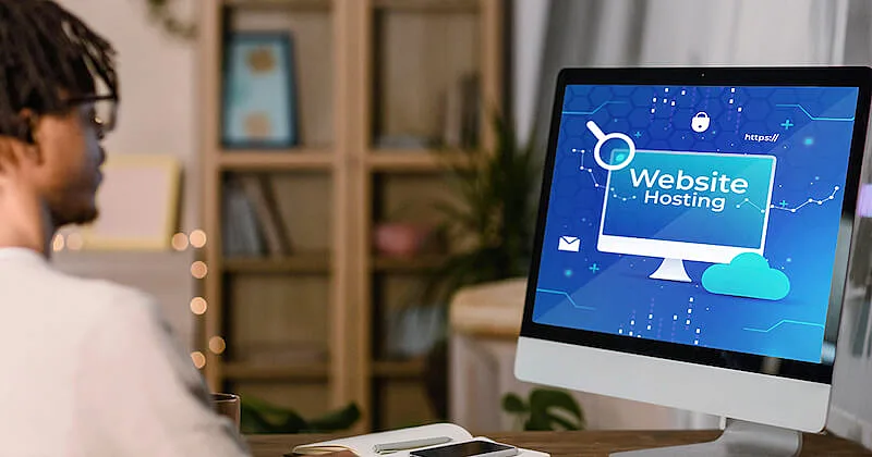 The image shows a man in front of a desktop computer. In the computer screen it's written "website hosting".