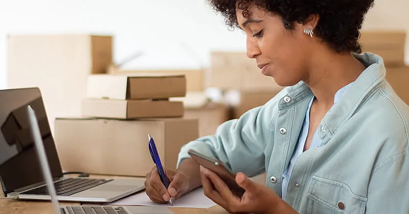 The image shows a woman in front of a laptop. She is wrting on a paper and holding a smartphone with the other hand. In the background there are several card boxes.
