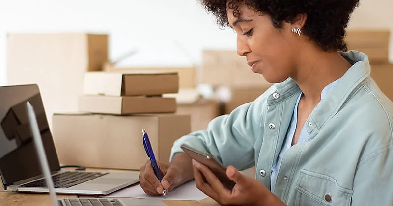 The image shows a woman in front of a laptop. She is wrting on a paper and holding a smartphone with the other hand. In the background there are several card boxes.