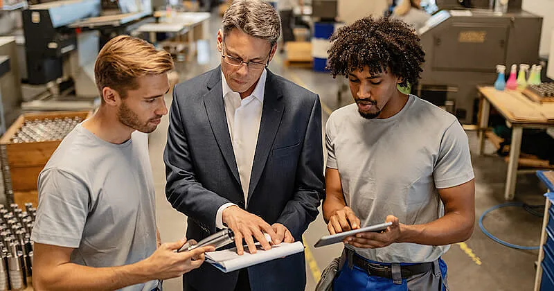 The image shows three man in a factory. they are holiding papers and analyzing them.