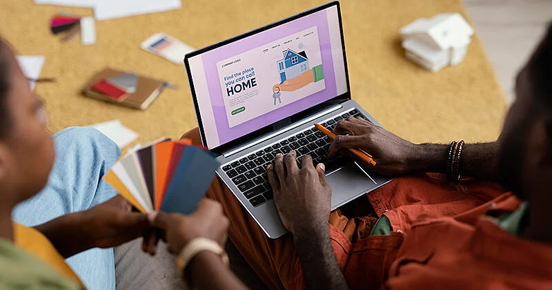 A person is in front of a laptop and the other one on their left side is holding a palette of colors. 