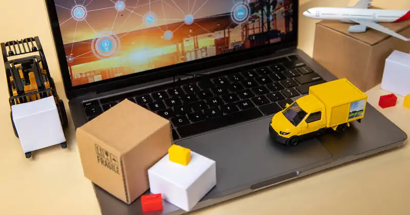 There is a laptop on a table and miniatures of a cruck, an airplane and cardboxes around it. 