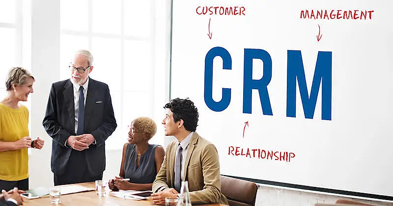 two women and two men are gathered in a meeting, in the white board behind them it's writtem "CRM. Customer relationship management".