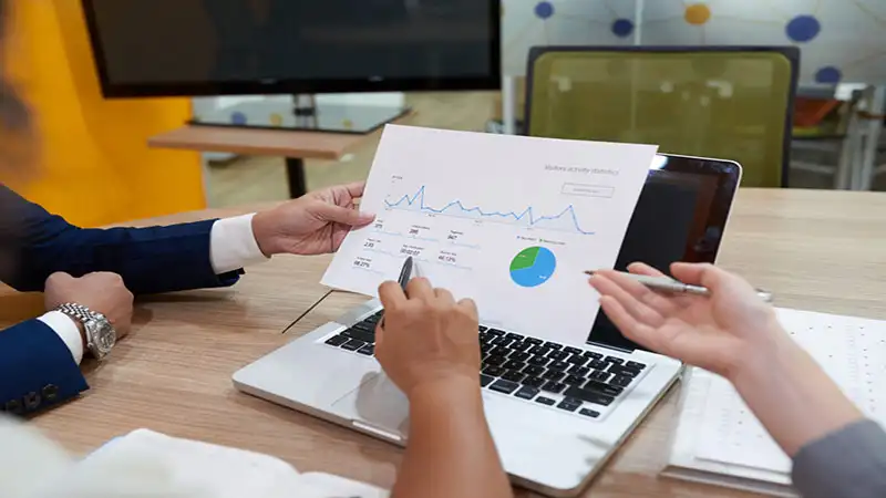 Foto de 3 pessoas analisando gráficos em uma folha e um notebook ao fundo
