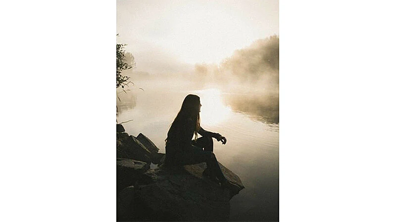 foto de uma mulher pensativa sentada em uma pedra na beira de um lago com neblina