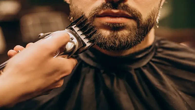 Foto de uma mão segurando um barbeador eletrico e passando na barba de um homem