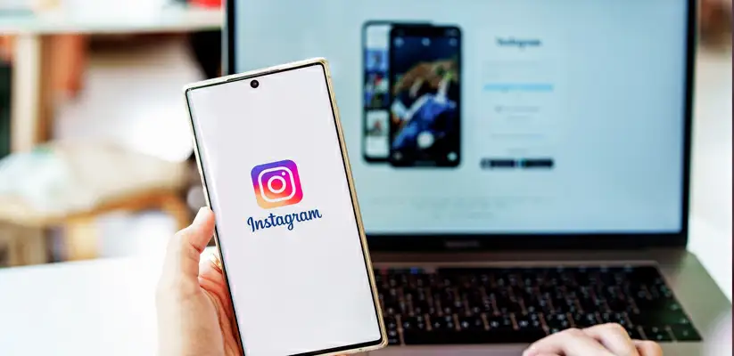 frases para divulgar uma loja no Instagram Foto de uma pessoa segurando o celular no Instagram e um notebook ao fundo