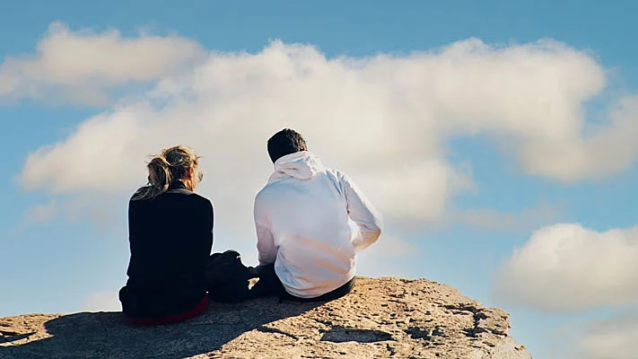 Foto de una pareja sentada al borde de las rocas, bajo un cielo azul