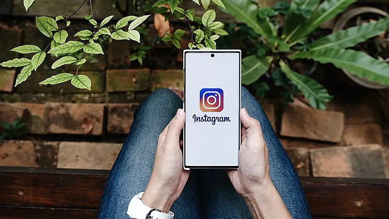 Uma foto de uma mão segurando um celular com o Instagram na tela e um notebook ao fundo com o Instagram aberto