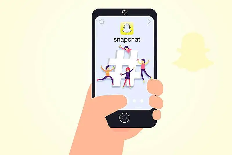 The image shows a person holding a smartphone. In the smarthphone there is the image of a hashtag with the Snapchat icon on the top of the screen.