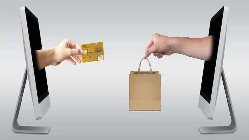 The image shows to computers inf front fo one another, in the one on the left side a hand is going out of the screen with a credit card, on the right site, a hand is going out of the other screen holding a paperbag