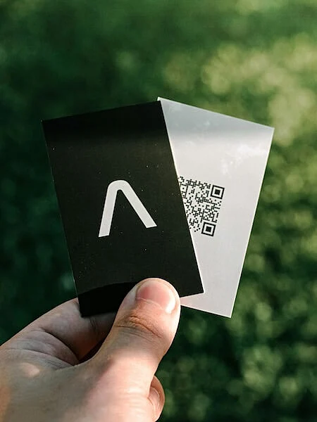 Person holding two minimalist digital business cards, one black with a white logo and one white with a printed QR code, against a blurred green background.