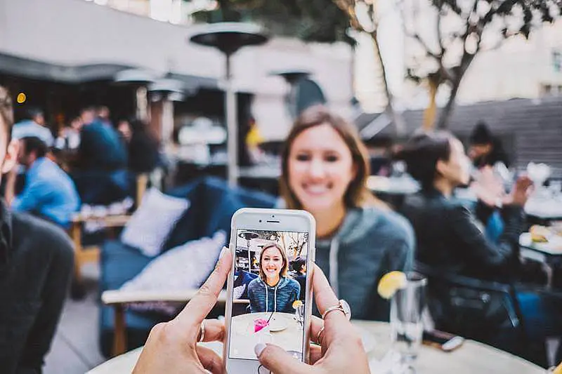 The image shows someone recording a girl. She is sitting on a table and smiling.