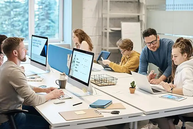 The image shows people working. They are sitting aroung a table, looking at their computers.
