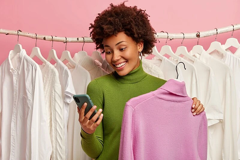 The image shows several clothes on a rack and a woman looking to a smartphone and holding a turtle neck shirt in fron of it.