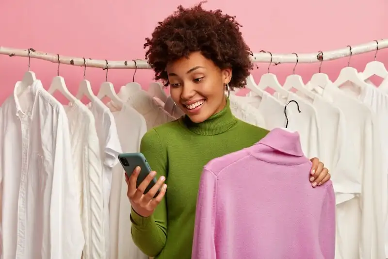 The image shows several clothes on a rack and a woman looking to a smartphone and holding a turtle neck shirt in fron of it.