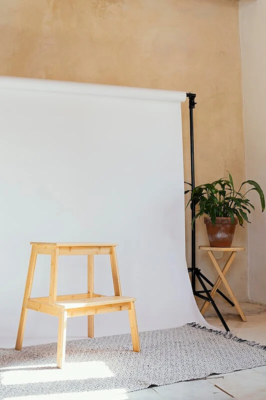 Whte backdrop, and in front of it there is a small stool, on the right side is a plant vase on a table. 