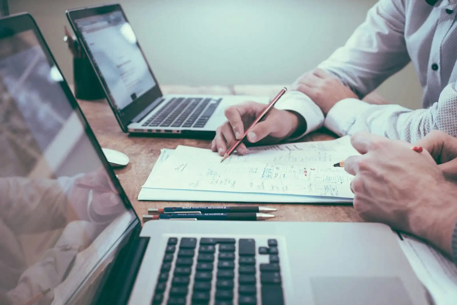 The image shows 2 laptos on a tables and two people in front of them, and looking at a paper