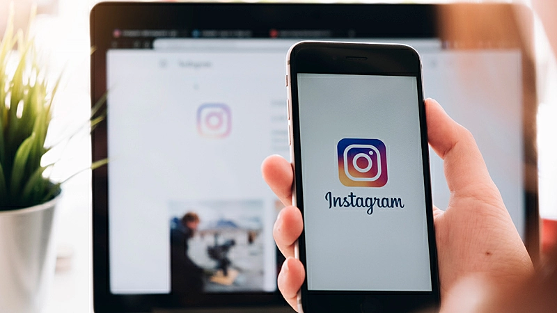 Foto de duas mão segurando o celular na tela de login do Instagram e um notebook ao fundo