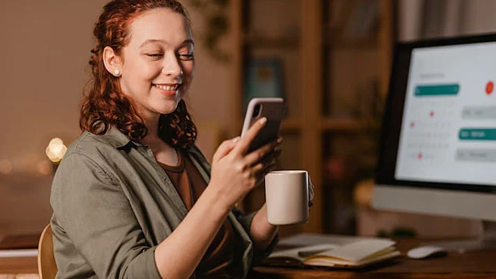 foto de una mujer usando su mobile frente a su computadora