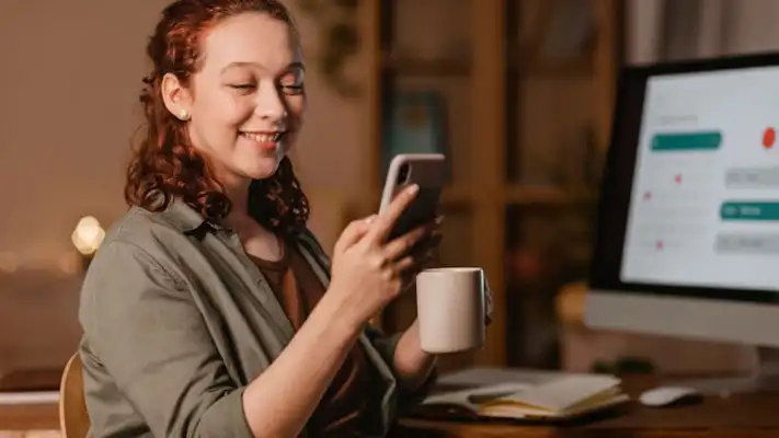 foto de una mujer usando su mobile frente a su computadora