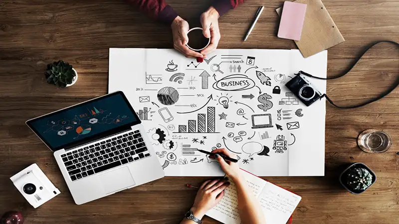 Foto duas pessoas trabalhando em uma mesa. Um laptop com gráficos, uma câmera, uma xícara de café e anotações visuais no centro, com desenhos e palavras relacionadas a estratégias empresariais.