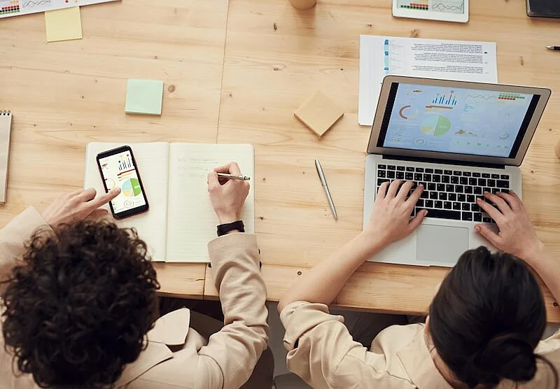 The image shows two men. The one in the left is viewing charts in a smartphone screen, and the one in the left is viewing charts in a laptop screen.