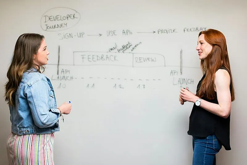 The image shows two women talking in fornt of a white board.