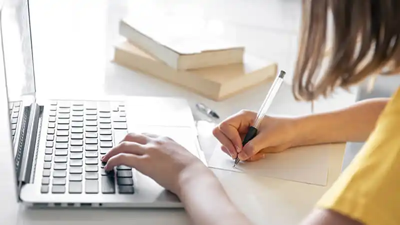 Foto de uma mulher fazendo anotações em um caderno e utilizando um notebook