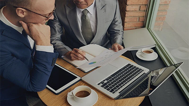 Foto de dois homens analisando um papel e um notebook em cima da mesa