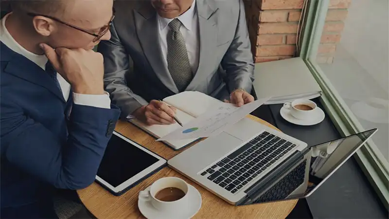 Foto de dois homens analisando um papel e um notebook em cima da mesa